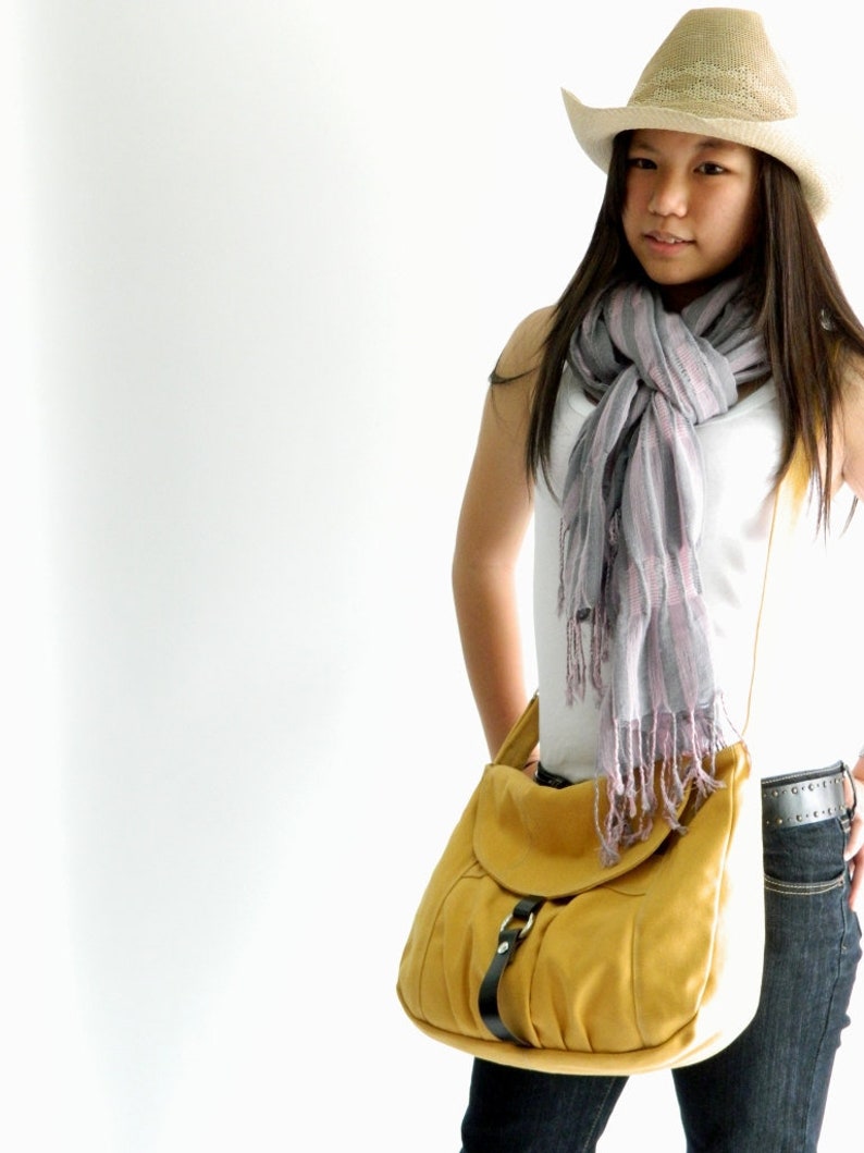 May include: A mustard yellow shoulder bag with a black buckle and strap. The bag is paired with a straw cowboy hat and a gray and pink scarf. The outfit is completed with dark wash jeans.