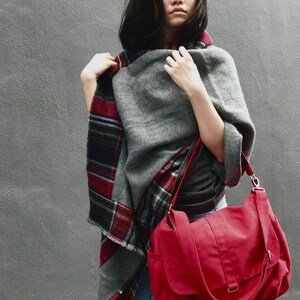 May include: A gray wool poncho with a red and black plaid border is worn with a red messenger bag. The bag has a flap closure and a shoulder strap. The background is a solid gray wall.