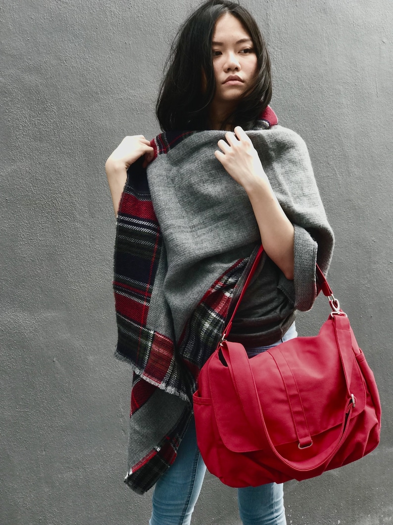 May include: A gray wool poncho with a red and black plaid border is worn with a red messenger bag. The bag has a flap closure and a shoulder strap. The background is a solid gray wall.