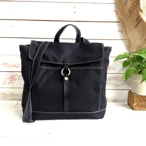 May include: A navy blue canvas backpack with white stitching details. The bag features a flap closure, a top handle, and a detachable shoulder strap. A black leather strap with a silver ring secures the flap. The bag is designed for everyday use.