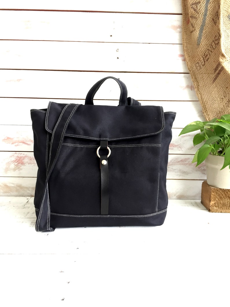May include: A navy blue canvas backpack with white stitching details. The bag features a flap closure, a top handle, and a detachable shoulder strap. A black leather strap with a silver ring secures the flap. The bag is designed for everyday use.