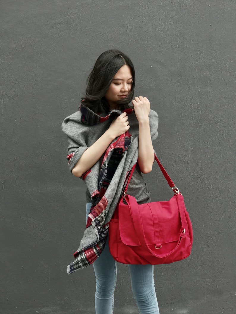 May include: A red canvas shoulder bag with a flap closure and adjustable strap. The bag is paired with a gray scarf with a red and black plaid pattern. The person is wearing a gray top and blue jeans.
