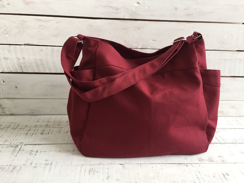 May include: A burgundy-colored tote bag with a shoulder strap and multiple pockets. The bag is made of a solid-colored fabric and has a simple, functional design. The background is a white wooden surface.