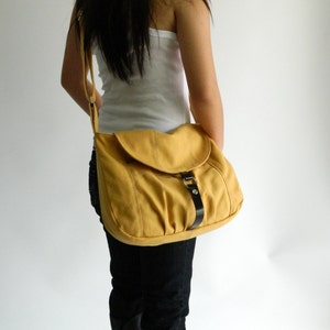 May include: Mustard yellow shoulder bag with a flap closure and a black leather strap. The bag is worn over the shoulder and has a buckle detail. The bag is made of a soft material.
