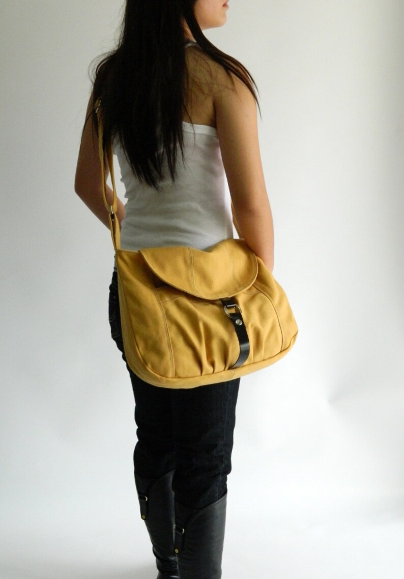 May include: Mustard yellow shoulder bag with a flap closure and a black leather strap. The bag is worn over the shoulder and has a buckle detail. The bag is made of a soft material.