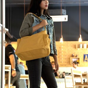 May include: A woman wearing a denim jacket and jeans is holding a large, mustard yellow tote bag with a brown strap. The bag is hanging from her shoulder and she is looking to the right.