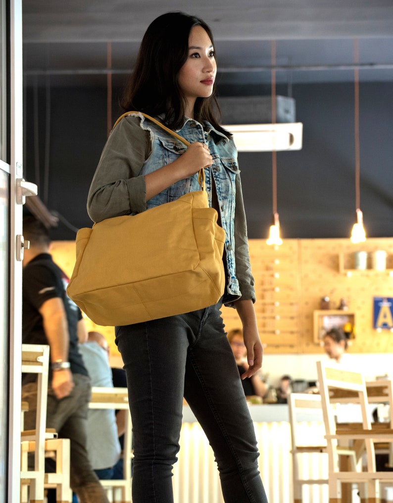 May include: A woman wearing a denim jacket and jeans is holding a large, mustard yellow tote bag with a brown strap. The bag is hanging from her shoulder and she is looking to the right.
