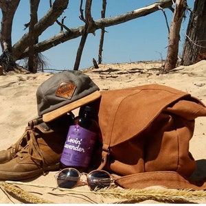 May include: A collection of outdoor accessories on a sandy beach. Includes a brown leather messenger bag, a gray cap with a "Twiddle" patch, brown boots, sunglasses, and a bottle of "Lovin' Lavender" tea.