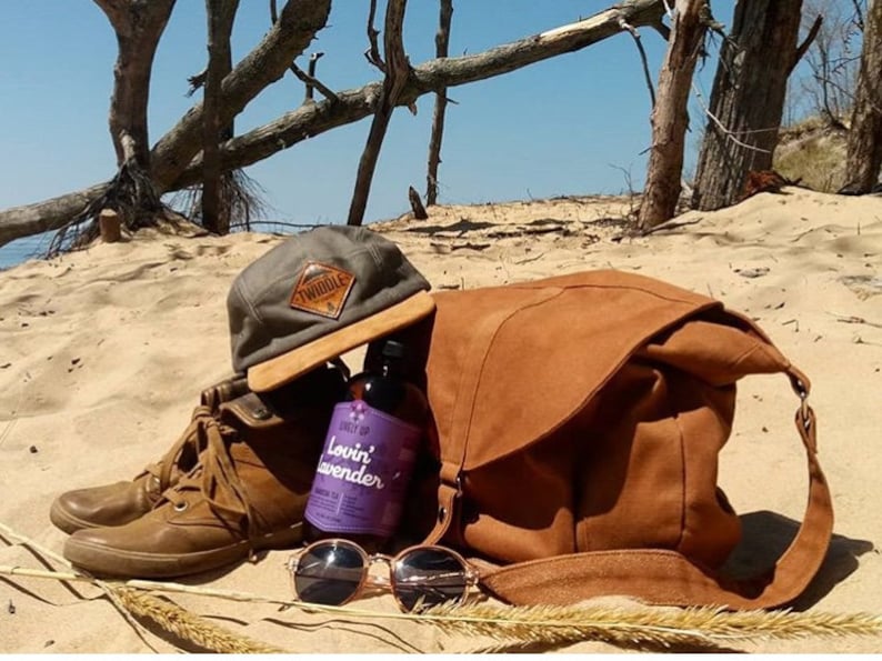 May include: A collection of outdoor accessories on a sandy beach. Includes a brown leather messenger bag, a gray cap with a "Twiddle" patch, brown boots, sunglasses, and a bottle of "Lovin' Lavender" tea.