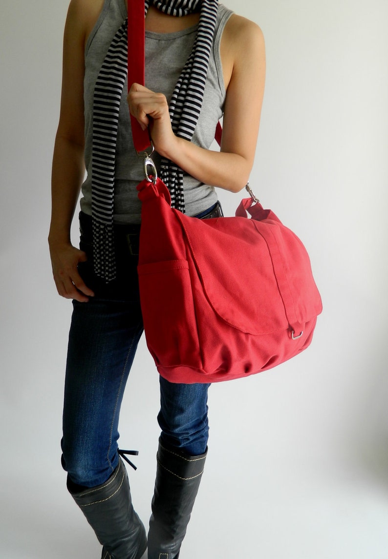 May include: A red canvas shoulder bag with a flap closure and a long, adjustable strap. The bag has a silver-colored metal clasp and a pocket on the front. The bag is being held by a person wearing a gray tank top, blue jeans, and black boots.