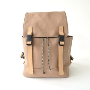 Puede incluir: Mochila de lona color canela con cierre de solapa y hebillas de plástico negro. La mochila presenta correas de cuero marrón claro y un cordón verde y blanco. Una bolsa elegante y funcional para el uso diario.