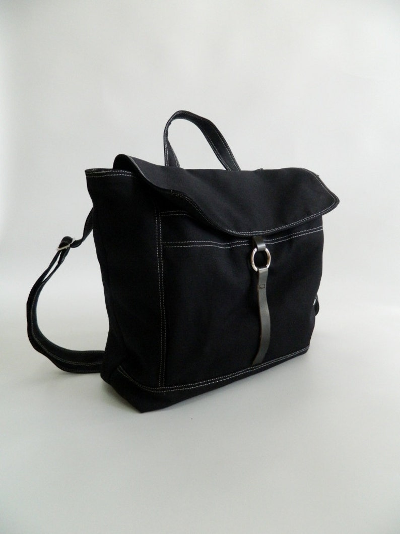 May include: A black backpack with a flap closure and a top handle. The bag features white stitching details and a leather strap with a metal ring. The straps are adjustable.