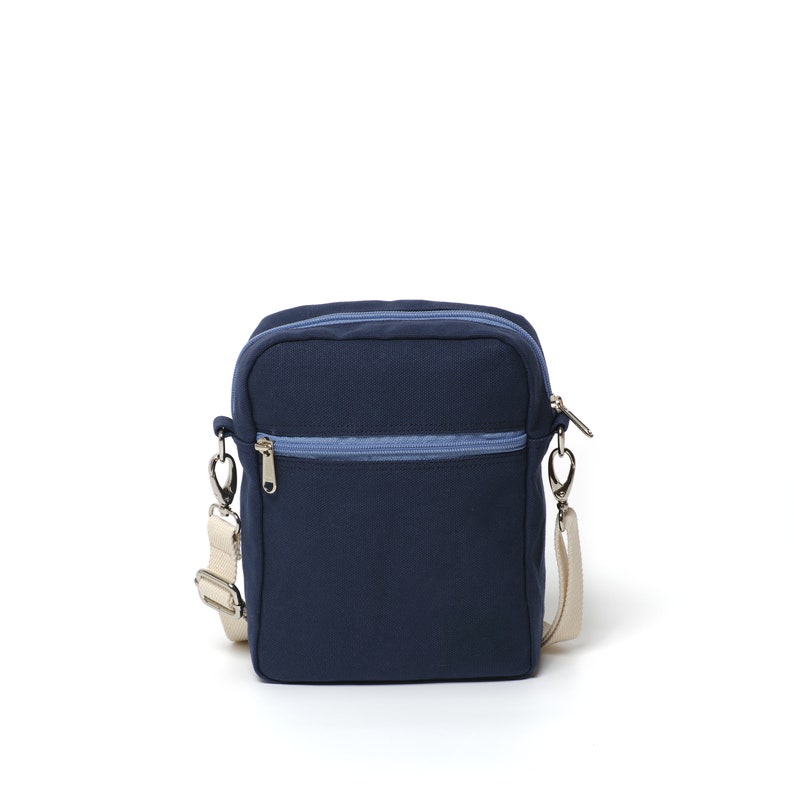 May include: A navy blue crossbody bag with a light blue zipper and a cream-colored adjustable strap. The bag has a rectangular shape and a silver-colored zipper pull. The bag is made of a durable fabric.