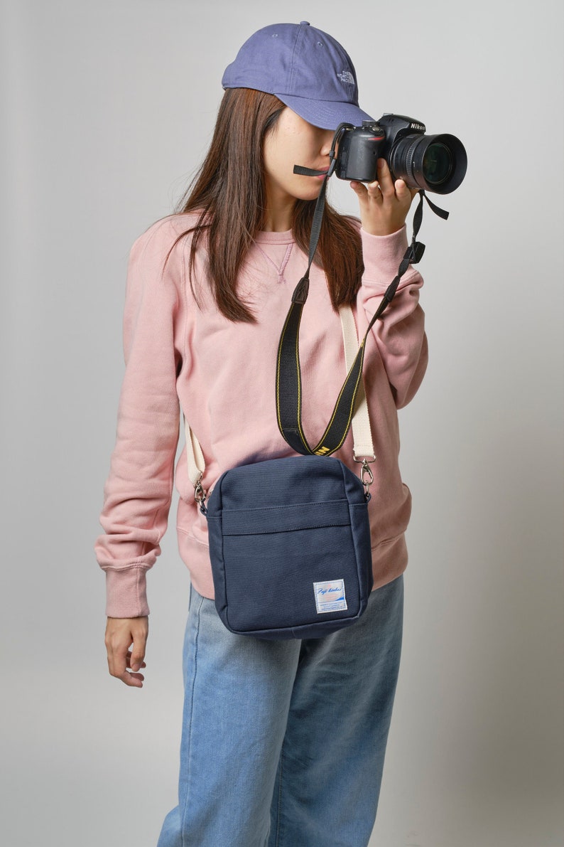 May include: A navy blue crossbody bag with a small white label. The bag has a rectangular shape and a long strap. The person in the image is wearing a pink sweatshirt, blue jeans, and a blue cap, and is holding a black camera.