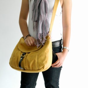 May include: Mustard yellow shoulder bag with a flap closure and black buckle detail. The bag has a long strap. A gray and pink striped scarf is draped around the neck.