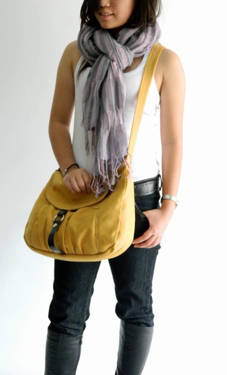 May include: Mustard yellow shoulder bag with a flap closure and black buckle detail. The bag has a long strap. A gray and pink striped scarf is draped around the neck.