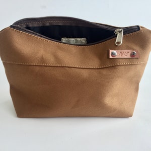 K&ouml;nnte beinhalten: Eine braune Canvas-Tasche mit Rei&szlig;verschluss, die ge&ouml;ffnet ist, um das dunkle Innere zu zeigen. Ein kleines Lederetikett mit dem Namen "IVIE" ist angebracht. Die Tasche hat eine rechteckige Form und ein schlichtes Design.