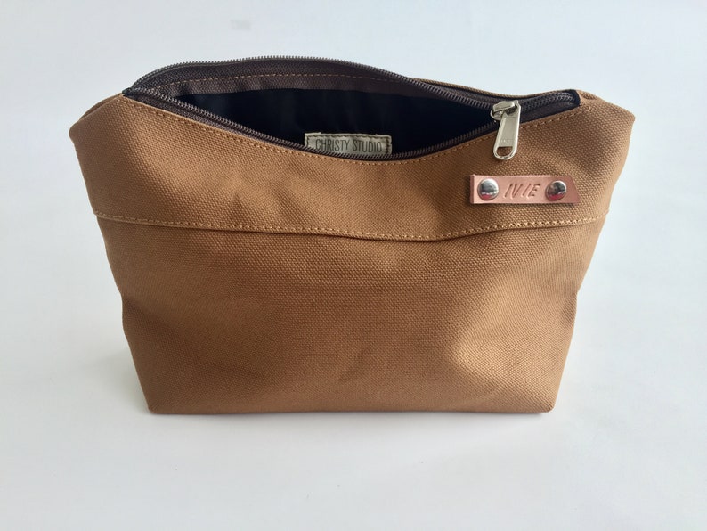 K&ouml;nnte beinhalten: Eine braune Canvas-Tasche mit Rei&szlig;verschluss, die ge&ouml;ffnet ist, um das dunkle Innere zu zeigen. Ein kleines Lederetikett mit dem Namen "IVIE" ist angebracht. Die Tasche hat eine rechteckige Form und ein schlichtes Design.