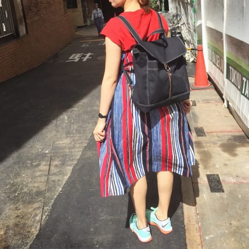 May include: A black backpack with a flap closure and a gold zipper is worn over a red top and a striped dress with red, white, and blue vertical stripes. The person is wearing blue and orange sneakers.