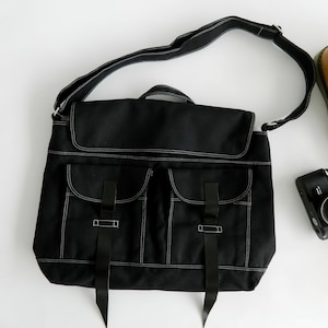 May include: Black messenger bag with white stitching, two front pockets, and a flap closure. The bag has a shoulder strap and two black straps with buckles. A camera and a pair of boots are also in the image.