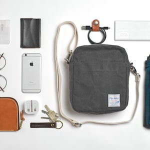 May include: An assortment of everyday carry items, including a gray crossbody bag, a smartphone, glasses, a wallet, keys, a power bank, and a compact umbrella. The items are arranged on a white surface.