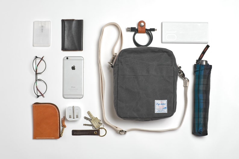 May include: An assortment of everyday carry items, including a gray crossbody bag, a smartphone, glasses, a wallet, keys, a power bank, and a compact umbrella. The items are arranged on a white surface.