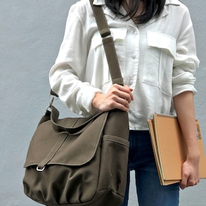 May include: Olive green canvas messenger bag with a flap closure, shoulder strap, and side pocket. The bag is carried by a person wearing a white button-down shirt and blue jeans. A spiral notebook is held in the other hand.
