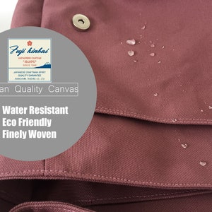 K&ouml;nnte beinhalten: Nahaufnahme einer staubrosa Canvas-Tasche mit Wassertropfen auf der Oberfl&auml;che. Die Tasche hat einen Klappenverschluss und einen Metallschnappverschluss. Der Text auf der Tasche lautet "Japan Quality Canvas", "Water Resistant", "Eco Friendly" und "Finely Woven".