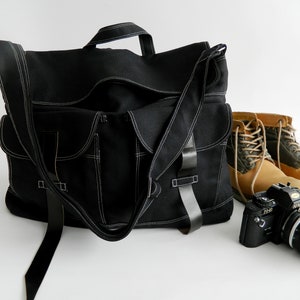 May include: Black messenger bag with white stitching, multiple pockets, and a shoulder strap. A pair of tan work boots and a black camera with a lens are next to the bag. The bag has a handle.