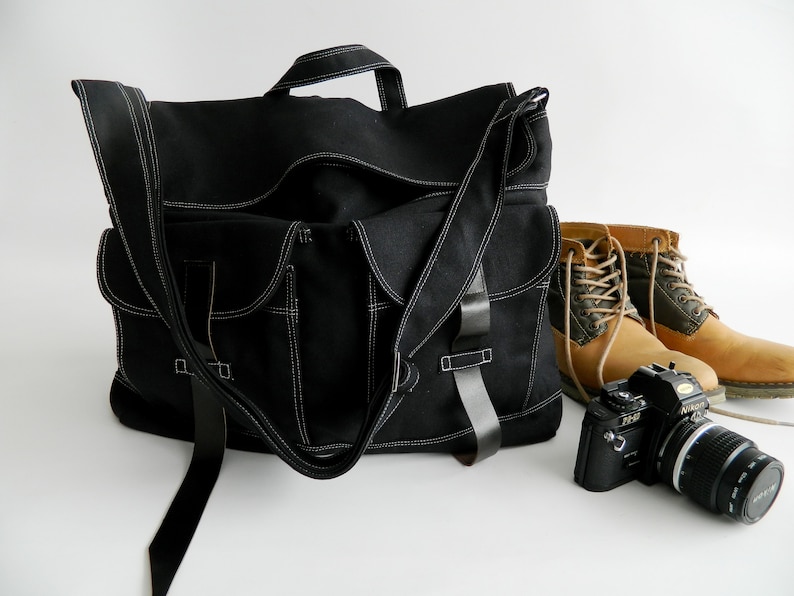 May include: Black messenger bag with white stitching, multiple pockets, and a shoulder strap. A pair of tan work boots and a black camera with a lens are next to the bag. The bag has a handle.