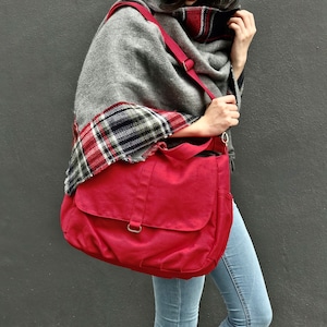 May include: A red messenger bag with a flap closure and a shoulder strap. The bag is carried over the shoulder. The person is wearing a gray shawl with a plaid pattern and light blue jeans.