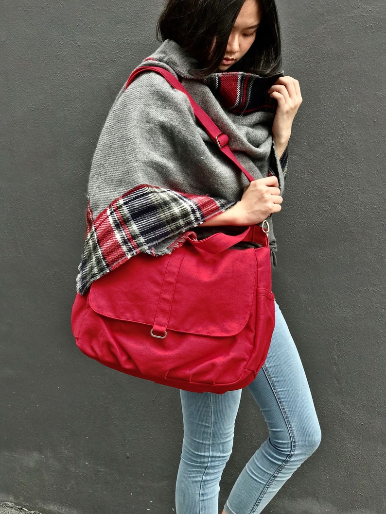 May include: A red messenger bag with a flap closure and a shoulder strap. The bag is carried over the shoulder. The person is wearing a gray shawl with a plaid pattern and light blue jeans.