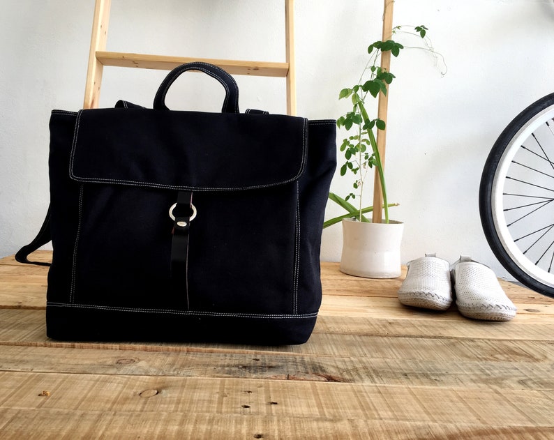 May include: A black canvas backpack with a flap closure and a top handle. The bag features white stitching and a leather strap with a silver ring closure. The bag is sitting on a wooden surface.