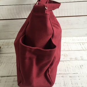 May include: A burgundy canvas tote bag with a shoulder strap and an exterior pocket. The bag is made of a sturdy material and has a simple, functional design. The bag is shown against a white wooden background.