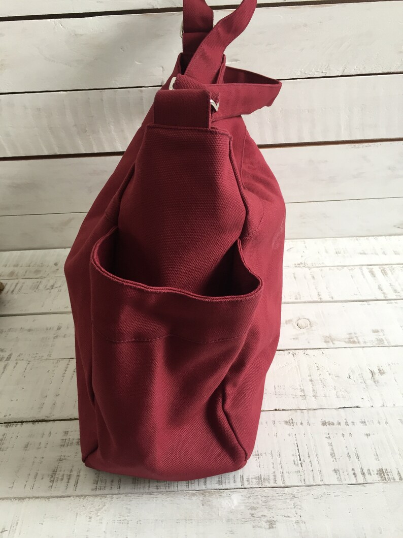 May include: A burgundy canvas tote bag with a shoulder strap and an exterior pocket. The bag is made of a sturdy material and has a simple, functional design. The bag is shown against a white wooden background.