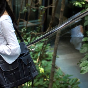 May include: Black messenger bag with two front pockets and a shoulder strap. The bag has white stitching and a flap closure. The bag is carried over the shoulder. The background includes greenery and a metal railing.