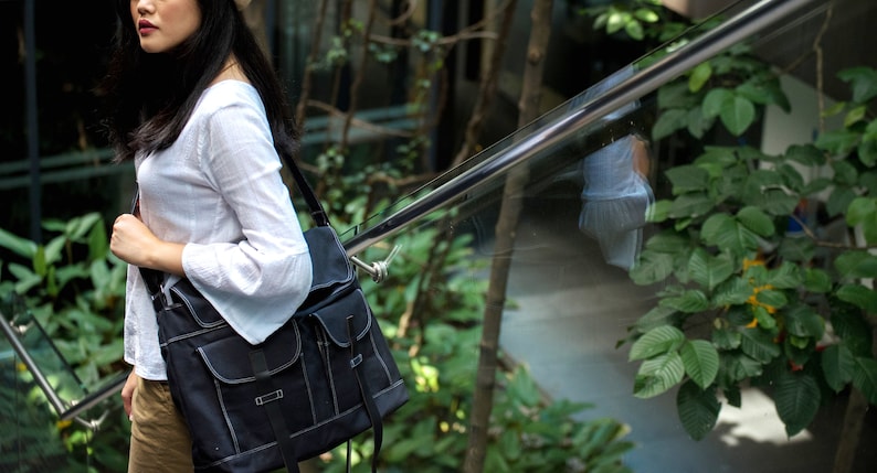 May include: Black messenger bag with two front pockets and a shoulder strap. The bag has white stitching and a flap closure. The bag is carried over the shoulder. The background includes greenery and a metal railing.