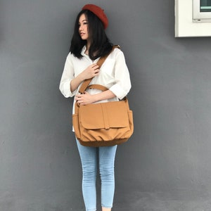 K&ouml;nnte beinhalten: Eine beige Messenger-Tasche mit Klappenverschluss und verstellbarem Schulterriemen. Die Person tr&auml;gt ein rostfarbenes Barett, ein wei&szlig;es Hemd, hellblaue Jeans und schwarze Stiefel.