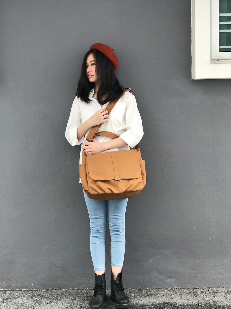 K&ouml;nnte beinhalten: Eine beige Messenger-Tasche mit Klappenverschluss und verstellbarem Schulterriemen. Die Person tr&auml;gt ein rostfarbenes Barett, ein wei&szlig;es Hemd, hellblaue Jeans und schwarze Stiefel.