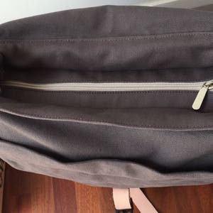May include: A dark gray canvas messenger bag with a zippered main compartment and multiple pockets. The bag features a light-colored zipper and a matching strap. The bag is designed for carrying items.
