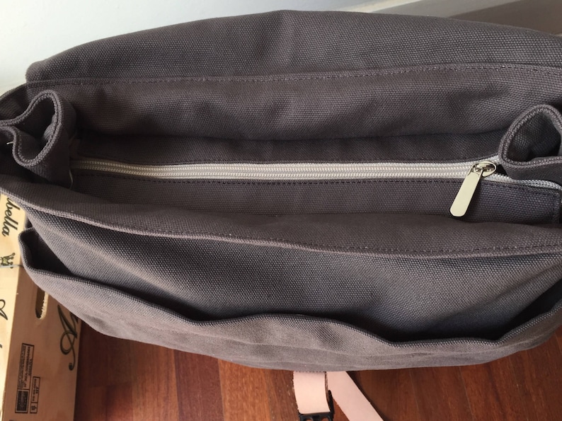 May include: A dark gray canvas messenger bag with a zippered main compartment and multiple pockets. The bag features a light-colored zipper and a matching strap. The bag is designed for carrying items.