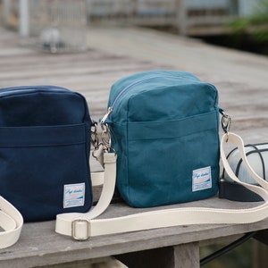 May include: Two canvas crossbody bags in navy blue and teal, each with a cream-colored adjustable strap. The bags have a rectangular shape with a front pocket and a small label. A black and white outdoor light fixture is also visible.
