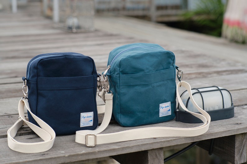 May include: Two canvas crossbody bags in navy blue and teal, each with a cream-colored adjustable strap. The bags have a rectangular shape with a front pocket and a small label. A black and white outdoor light fixture is also visible.