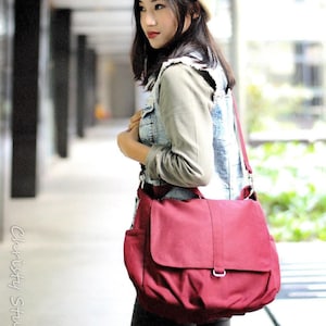 May include: A burgundy messenger bag with a flap closure and a shoulder strap. The bag has a rectangular shape with a pocket on the side. The bag is carried over the shoulder. The image also shows a person wearing a denim vest and a beige beret.
