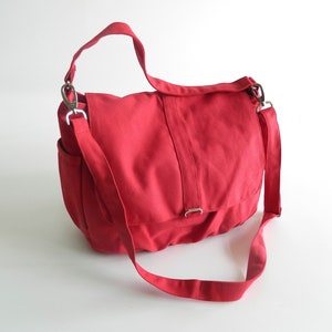 May include: A red canvas messenger bag with a flap closure and adjustable shoulder strap. The bag features a side pocket and silver-tone metal hardware. The bag is designed for carrying items and has a casual, everyday style.