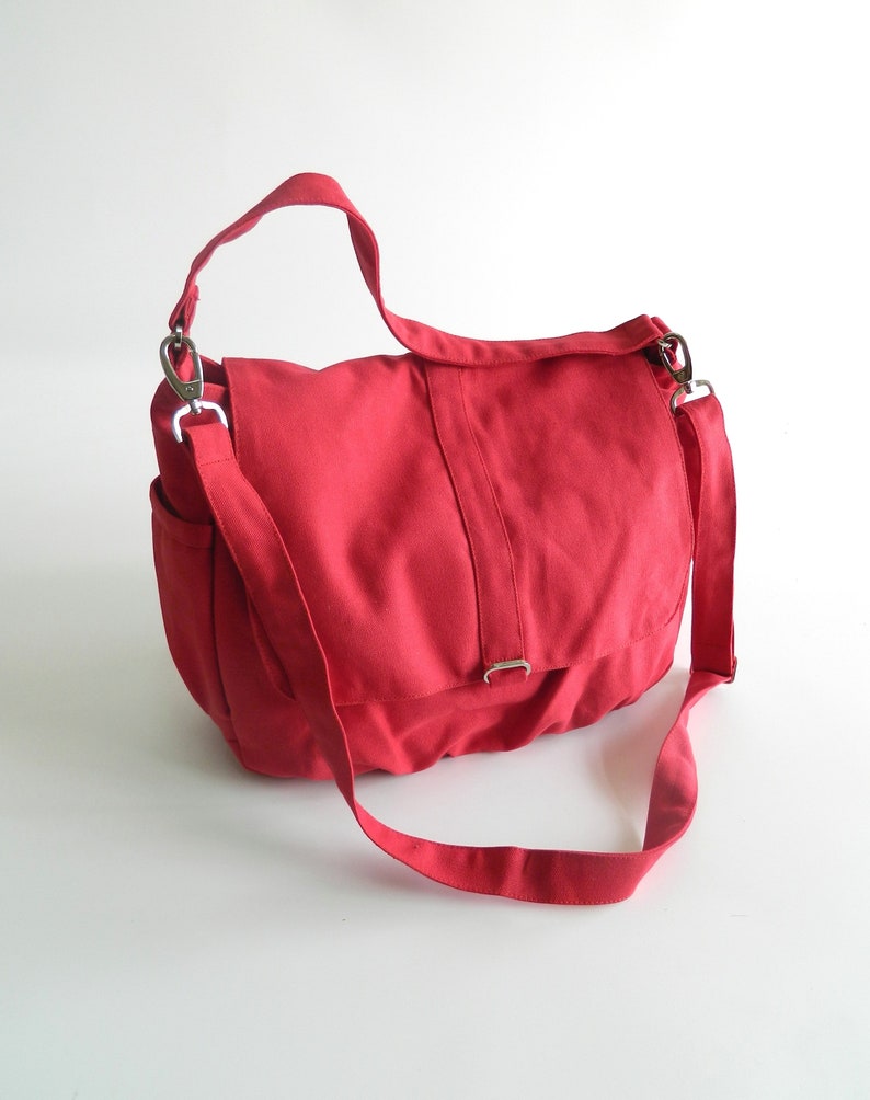 May include: A red canvas messenger bag with a flap closure and adjustable shoulder strap. The bag features a side pocket and silver-tone metal hardware. The bag is designed for carrying items and has a casual, everyday style.