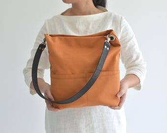 May include: An orange canvas tote bag with a black leather strap. The bag has two front pockets and silver-colored metal hardware. The bag is held in front of a person wearing a white shirt.