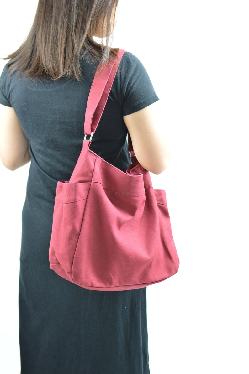 May include: A burgundy tote bag with a shoulder strap. The bag has two exterior pockets and a large main compartment. The strap is attached to the bag with silver metal rings. The bag is made of a sturdy fabric.