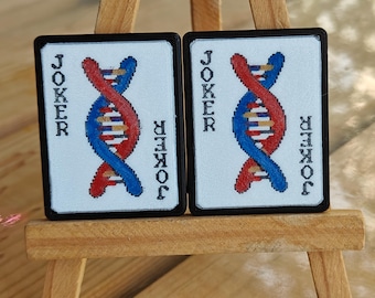 May include: Two playing cards with a white background, black borders, and the word "JOKER" on each side. The cards feature a DNA double helix design in red and blue. The cards are displayed on a small wooden easel.