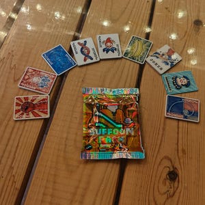 Puede incluir: Un conjunto de cartas de juego coloridas y un paquete envuelto en papel de aluminio están dispuestos sobre una superficie de madera. Las cartas presentan varios diseños, incluyendo un comodín, un plátano y otras ilustraciones únicas. El paquete tiene el texto "UFFOON".